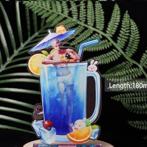 May include: A colorful acrylic standee featuring a man in a blue drink with a straw. The standee has a length of 180mm. It includes a lemon slice, ice cubes, and cartoon characters like a bunny and a bird. The background is a fern.