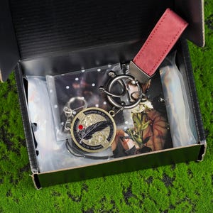 May include: A black gift box containing a keychain set. The set includes a round gold-coloured keychain with a feather design and the text "Everything is art", a red leather strap, and a silver-coloured metal keychain.
