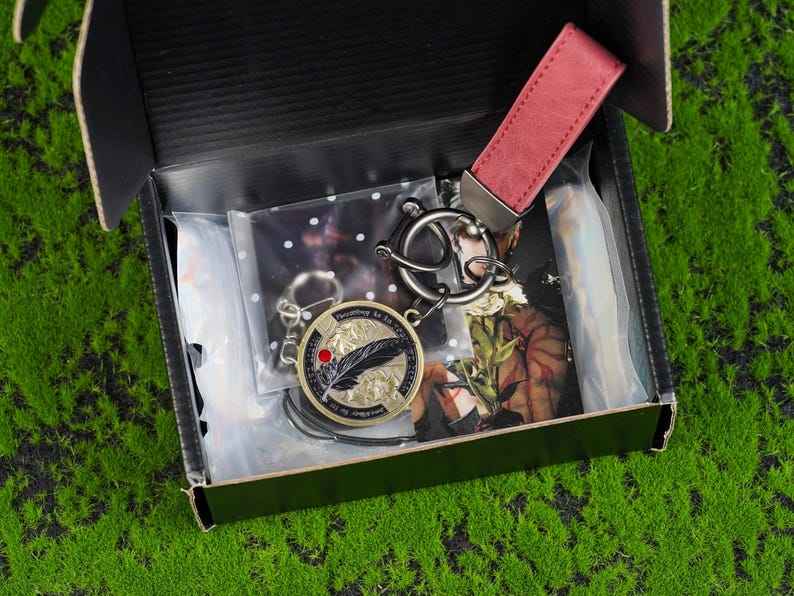 May include: A black gift box containing a keychain set. The set includes a round gold-coloured keychain with a feather design and the text "Everything is art", a red leather strap, and a silver-coloured metal keychain.