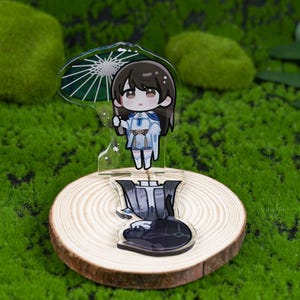 May include: A clear acrylic anime-style figure of a character holding an umbrella. The figure is on a wooden slice base. The character has brown hair and is wearing a blue and white outfit. The umbrella is green and white. The background is green.