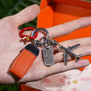 Könnte beinhalten: Eine Sammlung von Schlüsselanhängern zum Thema Reisen. Die Schlüsselanhänger umfassen ein orangefarbenes Lederetikett, einen silbernen Flugzeuganhänger und ein Etikett mit dem Text "When you come back". Die Schlüsselanhänger werden in einer Hand gehalten.