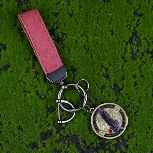 May include: A keyring with a burgundy leather strap and a silver-tone metal ring. The round pendant features a black feather design, a red dot, and text. The keyring is set against a green background.