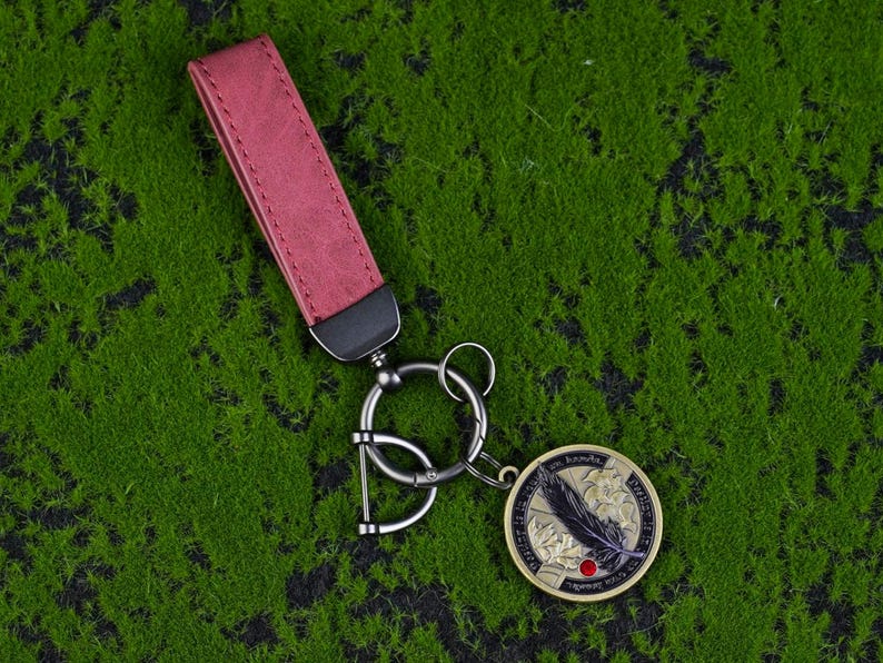 May include: A keyring with a burgundy leather strap and a silver-tone metal ring. The round pendant features a black feather design, a red dot, and text. The keyring is set against a green background.