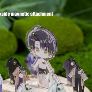 May include: A clear acrylic anime-style figure of a character with black hair and yellow eyes, with multiple outfits. The figure has a backside magnetic attachment. The background is a blurred green.