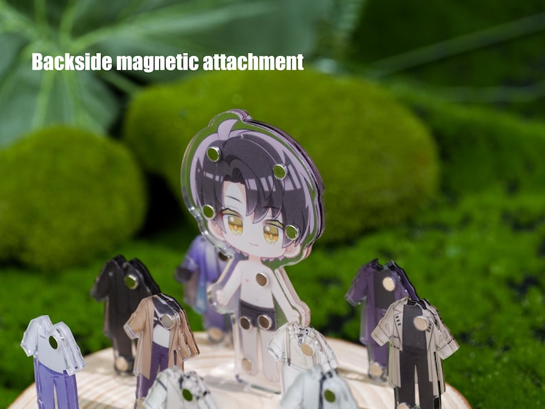 May include: A clear acrylic anime-style figure of a character with black hair and yellow eyes, with multiple outfits. The figure has a backside magnetic attachment. The background is a blurred green.