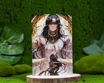 May include: A printed image of a character with long black hair and a blindfold, wearing ornate gold and red jewelry. The image is displayed on a wooden base against a backdrop of green moss and foliage.
