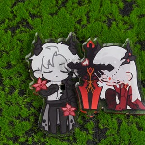 K&ouml;nnte beinhalten: Zwei Acryl-Anh&auml;nger mit Anime-Charakteren mit H&ouml;rnern und floralen Details. Ein Charakter ist in Schwarz und Wei&szlig;, der andere in Rot und Wei&szlig;. Ein schwarzes und rotes Schwert befindet sich zwischen ihnen. Die Anh&auml;nger befinden sich auf einer gr&uuml;nen, moosigen Oberfl&auml;che.