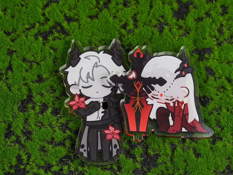 K&ouml;nnte beinhalten: Zwei Acryl-Anh&auml;nger mit Anime-Charakteren mit H&ouml;rnern und floralen Details. Ein Charakter ist in Schwarz und Wei&szlig;, der andere in Rot und Wei&szlig;. Ein schwarzes und rotes Schwert befindet sich zwischen ihnen. Die Anh&auml;nger befinden sich auf einer gr&uuml;nen, moosigen Oberfl&auml;che.
