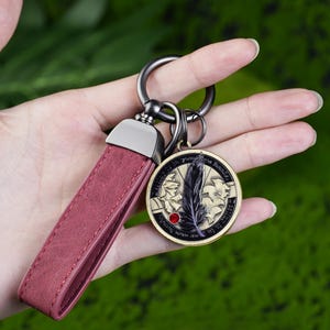 May include: A burgundy leather keychain with a metal clasp and a round pendant. The pendant features a black feather design, a red gem, and a gold-colored border. The keychain is held against a green background.