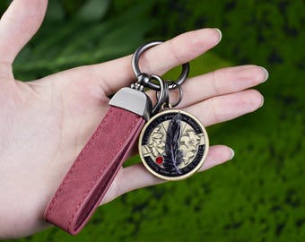 May include: A burgundy leather keychain with a metal clasp and a round pendant. The pendant features a black feather design, a red gem, and a gold-colored border. The keychain is held against a green background.