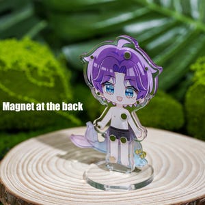 Peut inclure: Un charm en acrylique transparent repr&eacute;sentant un personnage de dessin anim&eacute; aux cheveux violets et aux yeux bleus. Le personnage est torse nu, porte un short noir et a une queue de sir&egrave;ne. Le charm est sur un support transparent. Le texte "Magnet at the back" est visible.