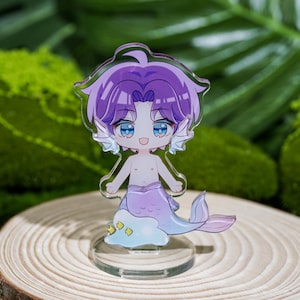 Peut inclure: Un charm en acrylique transparent repr&eacute;sentant un personnage de dessin anim&eacute; de sir&egrave;ne avec des cheveux violets et une queue lavande. La sir&egrave;ne a les yeux bleus et se tient sur une base transparente. Le charme est plac&eacute; sur un fond de mousse verte et une tranche de bois.
