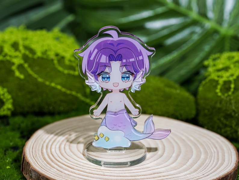 Peut inclure: Un charm en acrylique transparent repr&eacute;sentant un personnage de dessin anim&eacute; de sir&egrave;ne avec des cheveux violets et une queue lavande. La sir&egrave;ne a les yeux bleus et se tient sur une base transparente. Le charme est plac&eacute; sur un fond de mousse verte et une tranche de bois.