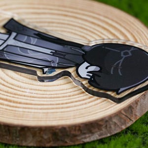 May include: A clear acrylic charm featuring a cartoon character with a black and gray outfit and a white glove. The charm is placed on a wooden slice, with a green background.