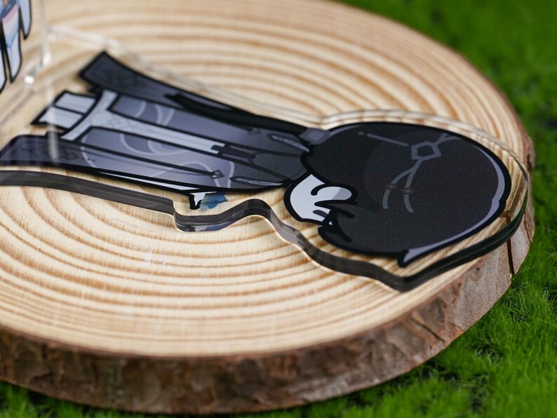 May include: A clear acrylic charm featuring a cartoon character with a black and gray outfit and a white glove. The charm is placed on a wooden slice, with a green background.