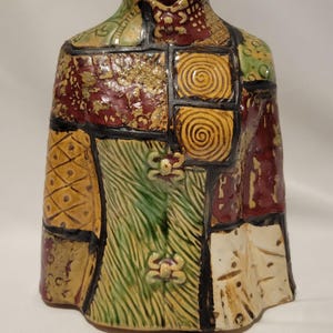 May include: A ceramic vase with a patchwork design. The vase features a variety of colors, including red, green, yellow, and brown. The design includes squares, spirals, and floral accents. The vase has a unique, textured surface.