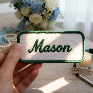 Op de afbeelding: Een wit rechthoekig embleem met een groene rand en de naam "Mason" geborduurd in groen cursief. Het embleem wordt in een hand gehouden, met een boeket witte en blauwe bloemen op de achtergrond.