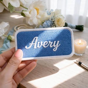 Pu&ograve; includere: Una toppa blu ricamata con il nome "Avery" in corsivo bianco. La toppa ha un bordo bianco ed &egrave; rettangolare con angoli arrotondati. Una candela accesa e dei fiori sono sullo sfondo.