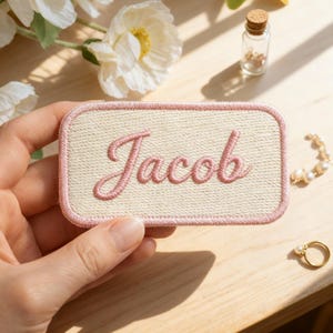 Peut inclure: Écusson rectangulaire avec le nom "Jacob" brodé en rose en écriture cursive. Le patch a un fond beige et une bordure rose. L'image comprend des fleurs et des bijoux, avec le patch tenu par une main.