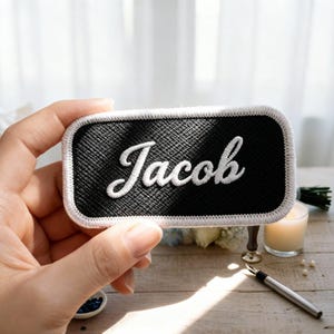 Pu&ograve; includere: Una toppa rettangolare nera con il nome "Jacob" ricamato in bianco. La toppa ha un bordo bianco. Lo sfondo include una candela, una penna e dei fiori. La toppa &egrave; tenuta in mano.