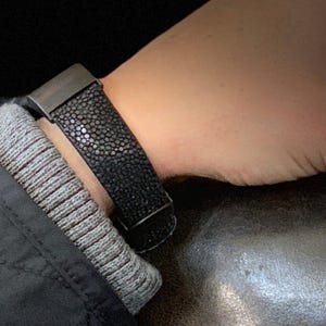 Op de afbeelding: Een zwart leren armband met een getextureerd oppervlak en een zilverkleurige sluiting. De armband wordt om een pols gedragen, met een donkere achtergrond. De armband lijkt van roggenleer te zijn.