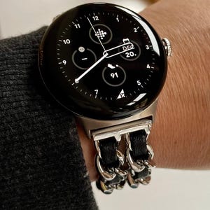 Può includere: Uno smartwatch nero con cinturino argentato e cinturino a catena in pelle nera. Il quadrante mostra l'ora, la data e altre informazioni. L'orologio è al polso.