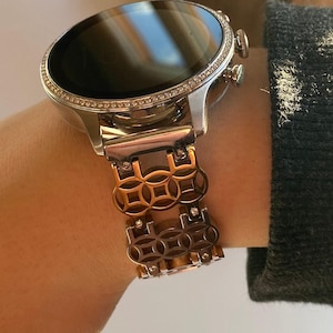 Könnte beinhalten: Eine silberne Smartwatch mit schwarzem Bildschirm und dekorativer Lünette. Die Uhr ist an einem kupferfarbenen Metallarmband mit geometrischem Design befestigt. Die Uhr befindet sich an einem Handgelenk.
