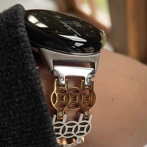 May include: A silver and gold-toned smartwatch with a black face and a decorative band. The band features a gold-colored geometric design with silver and diamond accents. The watch is worn on a wrist.