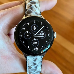 Könnte beinhalten: Eine schwarze Smartwatch mit goldfarbener Lünette und einem Armband mit Schlangenmuster. Das Zifferblatt zeigt die Uhrzeit, das Datum und andere Informationen an. Das Armband hat eine neutrale Farbe mit einem Schuppenmuster.
