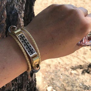 Puede incluir: Una pulsera de cuero dorado y beige con un diseño geométrico decorativo plateado y negro. La pulsera está envuelta alrededor de la muñeca. Unas gafas de sol teñidas de rosa se sostienen en la mano.