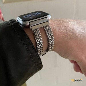 May include: Silver-toned watch band with a woven chain design, paired with a smartwatch. The band is secured with a clasp. The watch face displays the time. The watch and band are worn on a wrist.