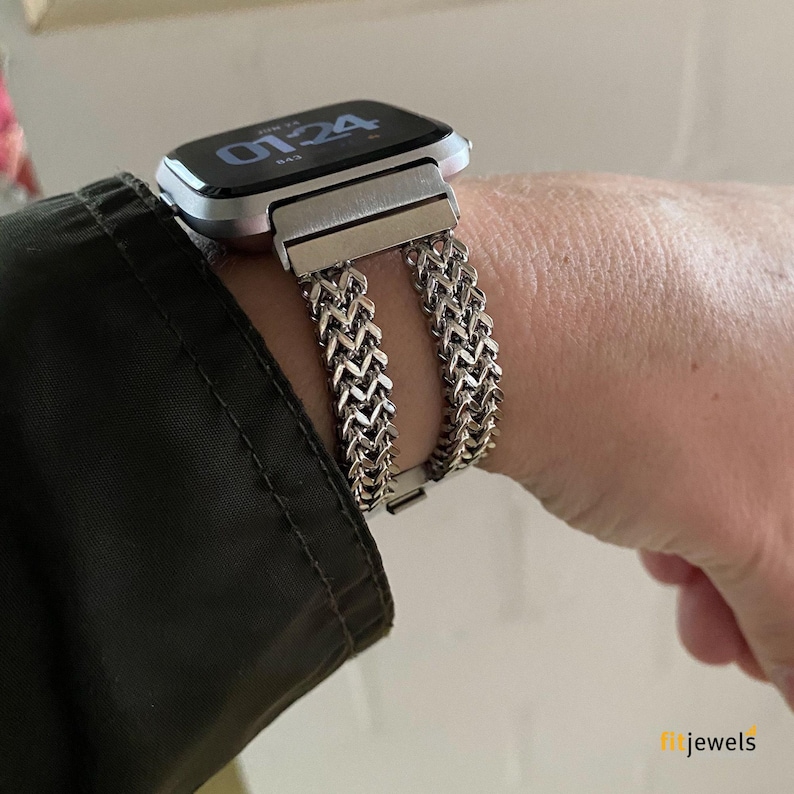 May include: Silver-toned watch band with a woven chain design, paired with a smartwatch. The band is secured with a clasp. The watch face displays the time. The watch and band are worn on a wrist.