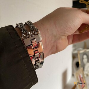 May include: A silver-toned bracelet with a chain and rectangular links. The bracelet is worn on a wrist, with a dark green jacket sleeve visible in the frame. The bracelet has a polished finish and a secure clasp.