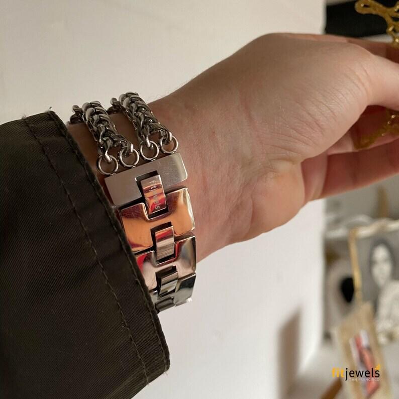 May include: A silver-toned bracelet with a chain and rectangular links. The bracelet is worn on a wrist, with a dark green jacket sleeve visible in the frame. The bracelet has a polished finish and a secure clasp.