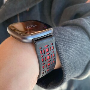 K&ouml;nnte beinhalten: Eine Smartwatch mit einem schwarz-roten Armband. Das Zifferblatt zeigt die Uhrzeit 12:25 Uhr an. Das Armband hat ein geometrisches Ausschnittmuster, das eine rote Unterschicht freilegt. Das Uhrengeh&auml;use ist dunkelmetallfarben.