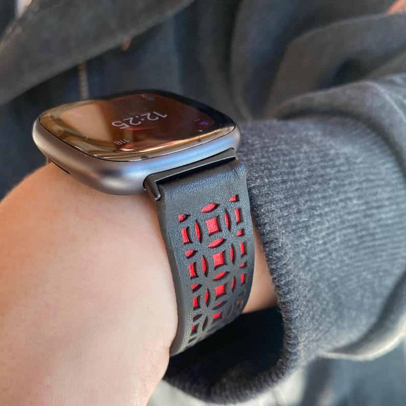 K&ouml;nnte beinhalten: Eine Smartwatch mit einem schwarz-roten Armband. Das Zifferblatt zeigt die Uhrzeit 12:25 Uhr an. Das Armband hat ein geometrisches Ausschnittmuster, das eine rote Unterschicht freilegt. Das Uhrengeh&auml;use ist dunkelmetallfarben.