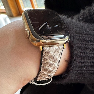 Könnte beinhalten: Eine goldfarbene Smartwatch mit schwarzem Bildschirm und einem Armband mit Schlangenmuster. Das Armband ist hellbraun und weiß. Die Uhr befindet sich an einem Handgelenk.