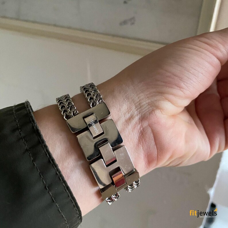 May include: A silver-toned bracelet with a unique design. The bracelet features a series of rectangular links connected by chain-like segments. The brand name "fitjewels" is visible on the bracelet. The bracelet is worn on a wrist.
