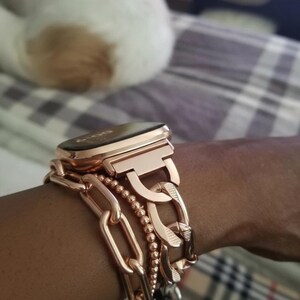 Może przedstawiać: Smartwatch w kolorze r&oacute;żowego złota z bransoletą z łańcuszka i bransoletką z koralik&oacute;w. Zegarek ma prostokątną tarczę i jest noszony na nadgarstku. Bransoletka składa się z dużych, zazębiających się ogniw. Bransoletka z koralik&oacute;w wykonana jest z małych, okrągłych koralik&oacute;w.