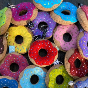 May include: A collection of felt donut keychains in various colors, including blue, green, yellow, pink, red, and purple. Each donut has colorful sprinkles and a brown felt base.