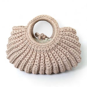 May include: A light pink, hand-crocheted handbag with a circular handle. The bag has a rounded, fan-like shape with a drawstring closure visible inside. The thick yarn creates a textured, sturdy appearance. The bag is set against a white background.