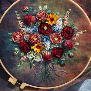 Può includere: Ricamo di un bouquet floreale in un cerchio di legno. Il bouquet presenta fiori rossi, gialli e blu con foglie verdi. Il tessuto di sfondo è un mix di colori. Il testo in basso recita "Message for custom".
