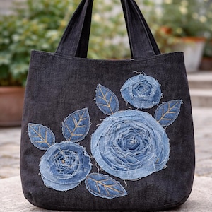 Puede incluir: Un bolso tote de mezclilla negra con un diseño floral. El bolso presenta tres grandes rosas de mezclilla azul y varias hojas de mezclilla azul con costuras doradas. El bolso tiene dos asas negras.