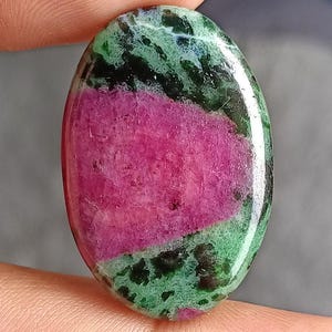 Peut inclure: Pierre précieuse de forme ovale, présentant une combinaison de couleurs vives. La pierre affiche une bande proéminente de rose foncé ou magenta, contrastant avec des zones vertes et noires. La surface polie reflète la lumière.