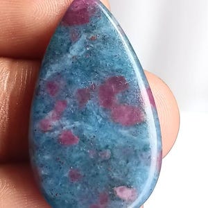 Op de afbeelding: Een druppelvormige, gepolijste edelsteen. De steen heeft een mix van blauwe en roze tinten, met het roze in onregelmatige vlekken over de blauwe basis. De edelsteen wordt door een hand vastgehouden.
