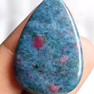 Puede incluir: Una gema pulida en forma de lágrima. La piedra presenta un patrón moteado de tonos azul-verdosos, salpicado de parches rosados vibrantes. La gema se sostiene en una mano, resaltando su superficie lisa y reflectante.