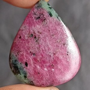 Könnte beinhalten: Ein polierter, tropfenförmiger Edelstein mit einem lebendigen rosa-grünen Farbschema. Der Stein weist eine Mischung aus rosa und grünen Farbtönen mit dunklen Sprenkeln auf, wodurch ein einzigartiges und natürliches Muster entsteht. Der Edelstein wird zwischen zwei Fingern gehalten.