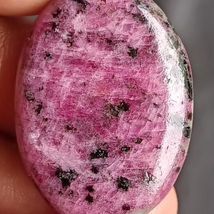 Könnte beinhalten: Ein polierter, oval geformter Edelstein in leuchtendem Rosa, gesprenkelt mit dunklen schwarzen Einschlüssen. Der Stein wird zwischen den Fingern gehalten, wodurch seine glatte, reflektierende Oberfläche hervorgehoben wird. Die Farbe und das Muster des Edelsteins erzeugen einen auffälligen visuellen Kontrast.