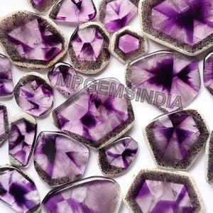 Può includere: Una collezione di pietre preziose di ametista lucidata in varie forme. Le pietre mostrano una gamma di tonalità viola con motivi interni e un bordo bianco e nero. Il testo "MPGEMSINDIA" è visibile.