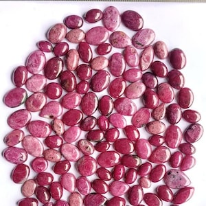 Pode incluir: Uma coleção de pedras preciosas ovais em tons de rosa e vermelho, dispostas sobre uma superfície branca. As pedras polidas variam em tamanho e tonalidade. Uma régua metálica, com a inscrição "MADE IN INDIA", é visível no fundo.